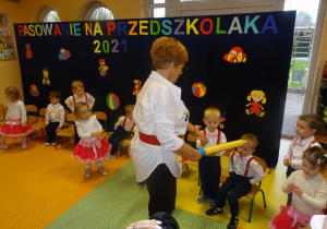 Pani dyrektor Maria Królikowska trzyma w ręku żółtą kredkę, wyciąga lewą dłoń do chłopca, który siedzi na krześle.